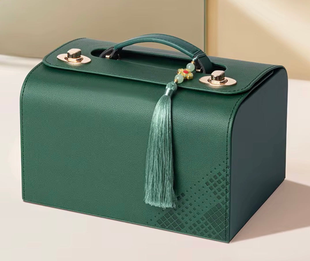 A structured dark green leather cosmetic case with a top handle, silver-tone hardware, a decorative tassel, and subtle geometric pattern on the lower front, photographed on a clean beige studio background.