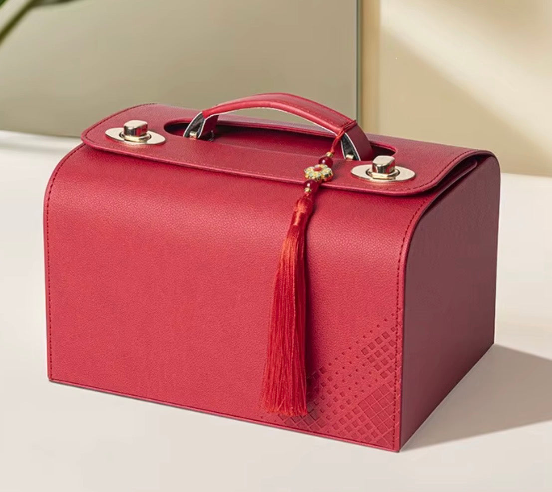 A red faux-leather cosmetic case with a top handle, silver-tone snap closures, and a red tassel charm, shown on a light beige surface.