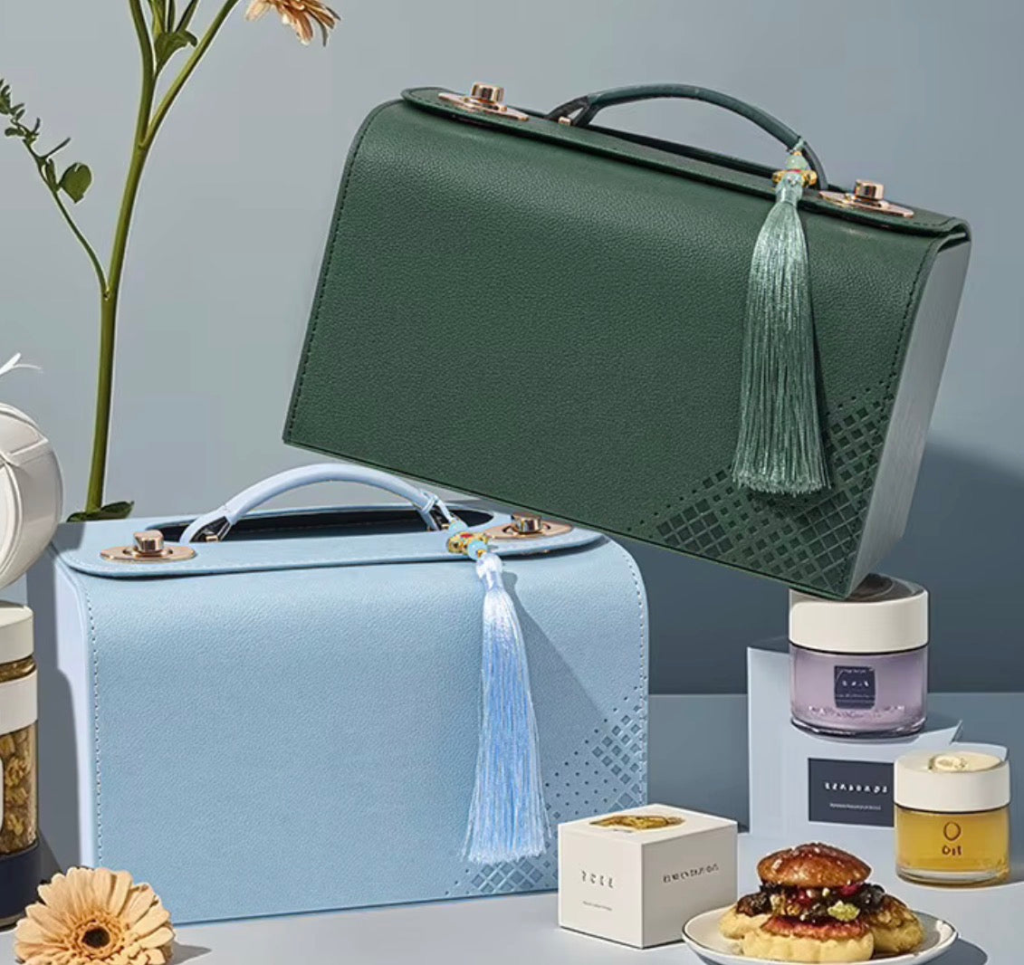 Two structured leather handbags are displayed in a styled studio setting, one in dark green and one in light blue, with decorative flowers, pastries, and small cosmetic containers around them.