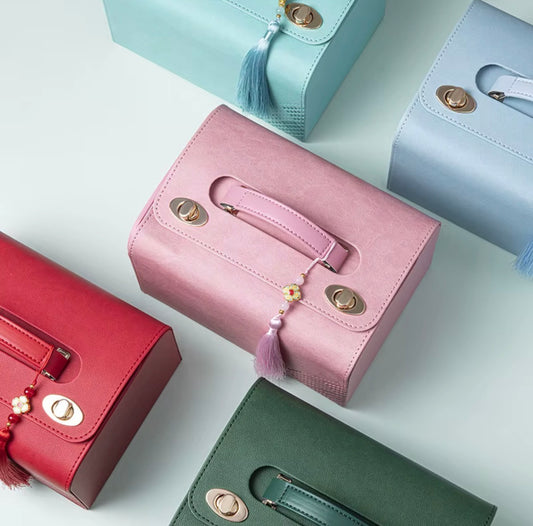 Five structured handbags in pastel and bright colors are arranged on a clean white background. The center bag is dusty pink with a tassel charm and gold-tone hardware, surrounded by red, mint, light blue, and dark green versions of the same bag style.
