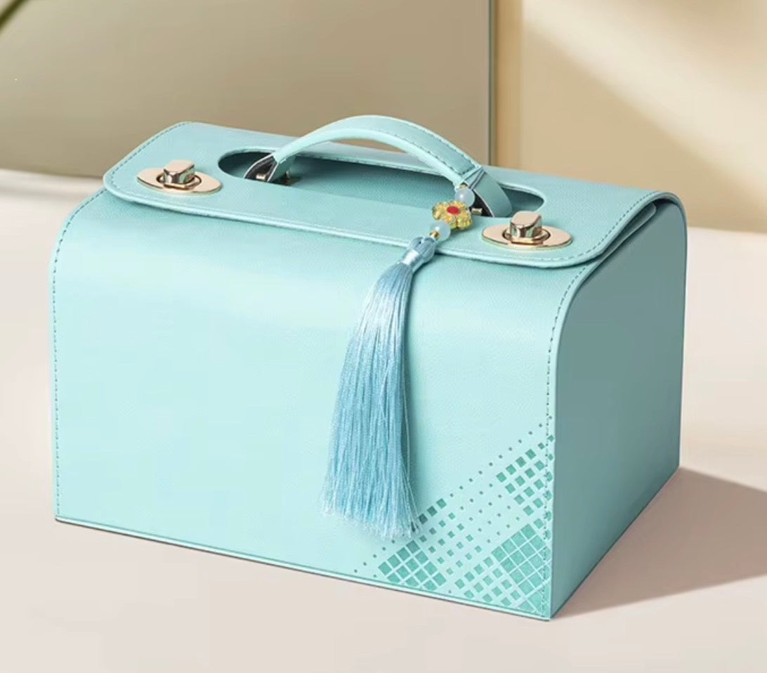 A light blue faux leather cosmetic case with a top handle, gold-tone hardware, a decorative blue tassel charm, and a subtle geometric pattern on the lower front, shown on a neutral beige background.