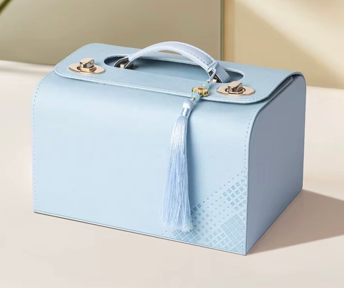 A light blue structured handbag with a top handle, gold-tone hardware, and a matching tassel charm, photographed on a warm beige surface.