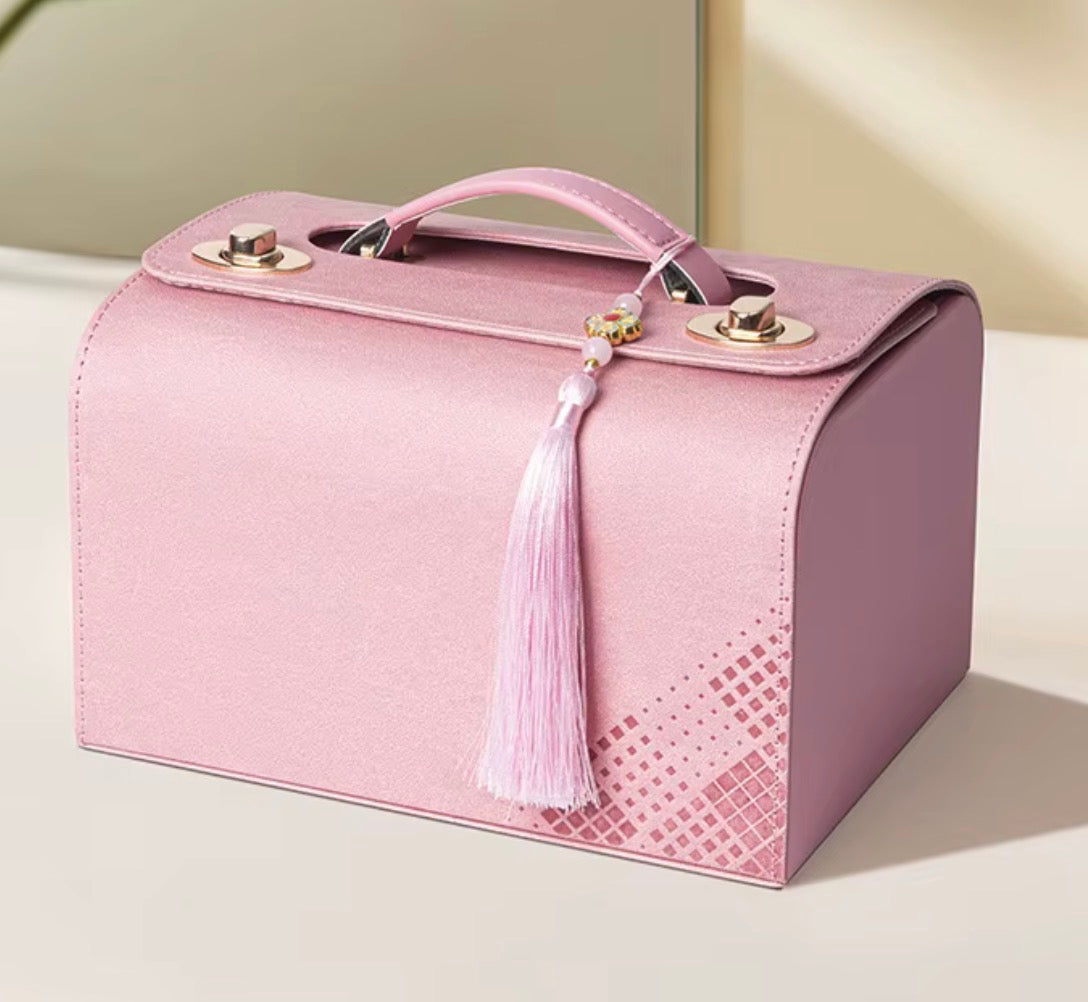 A pink structured cosmetic case or small handbag with a top handle, gold-tone hardware, and a hanging tassel, photographed on a clean light beige surface.