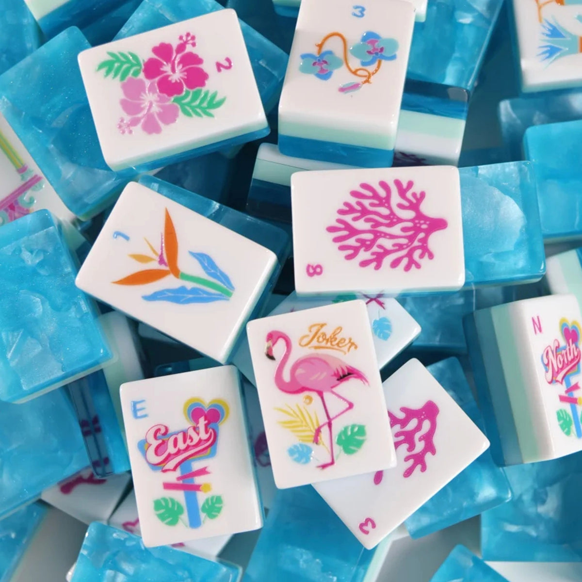A pile of square white resin tiles with colorful tropical and animal illustrations, including hibiscus flowers, a flamingo, a bird of paradise, and coral, mixed with translucent blue cubes.