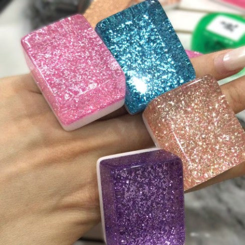 Four glitter tile color samples in pink, blue, peach, and purple, held in a hand.
